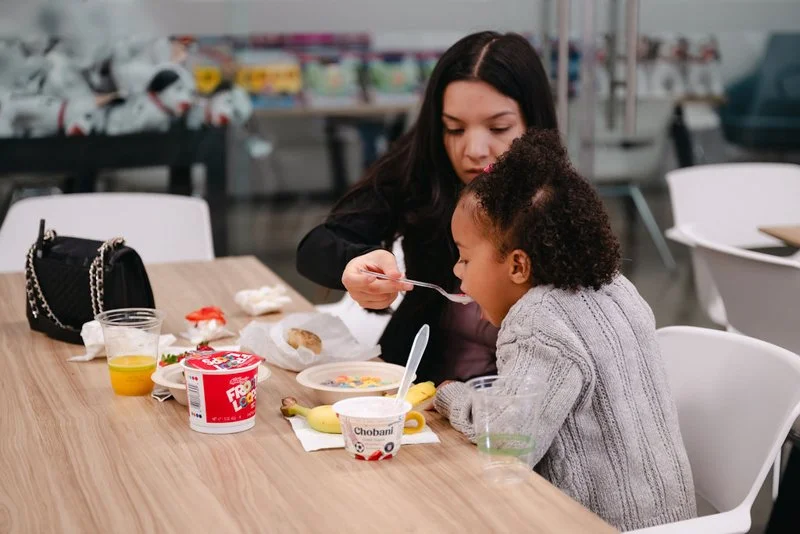 A woman feeding a young girl breakfast at a table in a casual dining area.