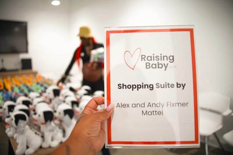 A person holding a sign that says 'Raising Baby, Shopping Suite by Alex and Andy Fixmer, Mattel' in front of a display of plush toys.