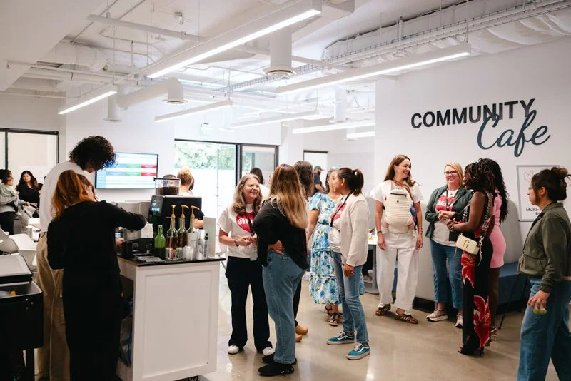 People gathered at a community cafe, socializing and chatting. The cafe has modern decor with white walls and large windows, and a coffee station with baristas serving drinks.