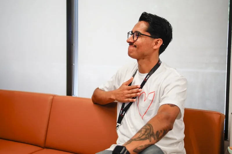 A man with glasses, tattoos, and a white t-shirt with a red heart, is sitting on an orange couch, holding his chest with his right hand, and appears to be in a thoughtful or emotional moment.