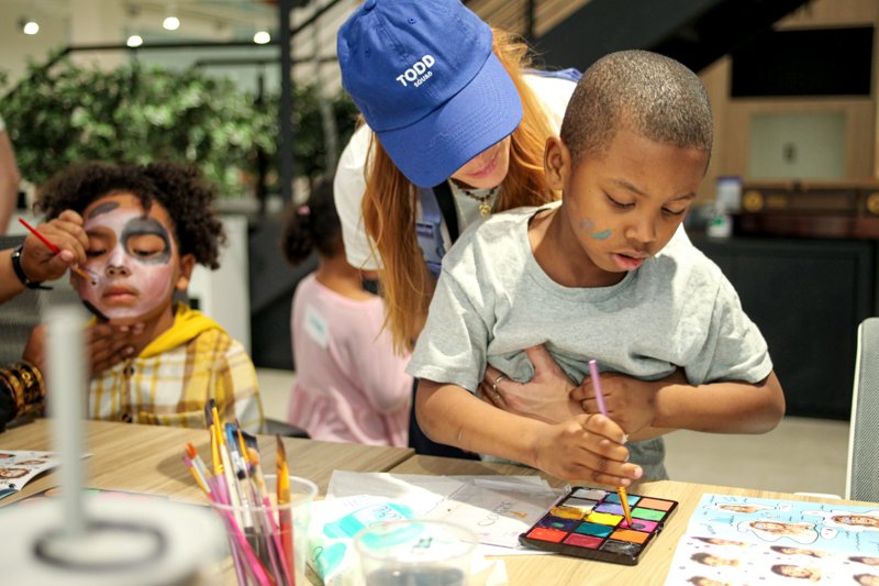 A young boy with face paint working on a colorful art project at a table, with a woman assisting him, and another child getting face painted in the background.