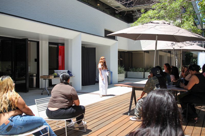 A woman with long red hair standing on a wooden deck speaking to a group of people seated outside under umbrellas.