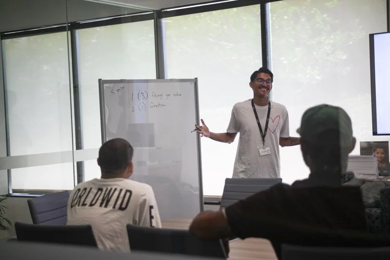 A man standing in front of a whiteboard and presenting to a small group in a modern conference room with large windows, while two people sit at the table facing him.