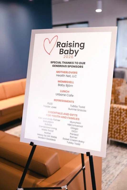 Signboard for a 2026 event called 'Raising Baby' with sponsors and refreshments listed, placed on a black stand indoors.