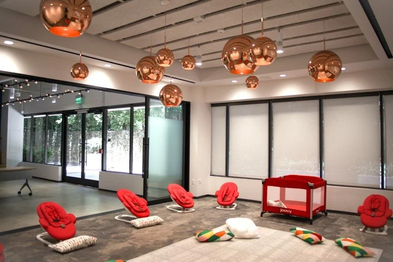 Modern lounge with hanging copper-colored spherical pendant lights, large windows with blinds, red chairs with swivel bases arranged in a semi-circle, a red crib-like structure, and colorful plush toys on the floor.