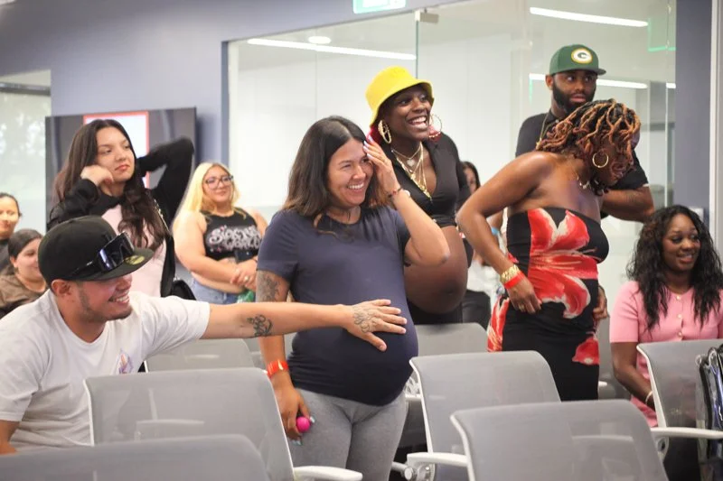 Group of diverse people in a room, some are standing and some are sitting, appearing to be engaged in a joyful or celebratory activity.