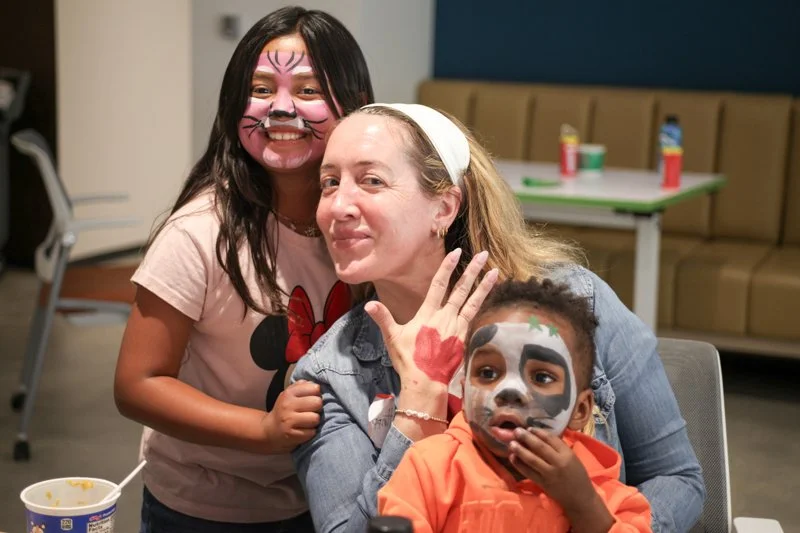 A woman with face paint and two children with face paint, one girl with face paint and a boy with zombie face paint, smiling and holding hands at an indoor event.