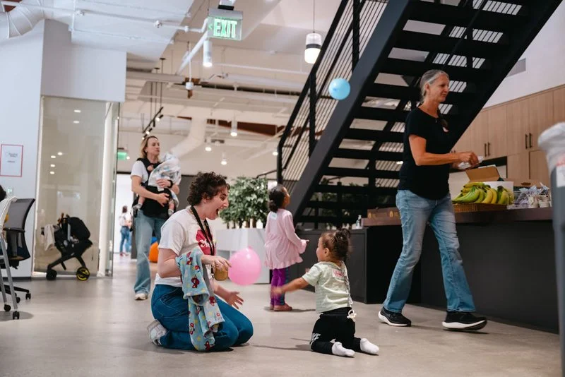 A woman kneeling on the floor holding a pink balloon and playing with a seated young girl in a yellow shirt. Two other children and a woman holding a baby are visible in the background inside a modern office or lobby space with a staircase and wooden