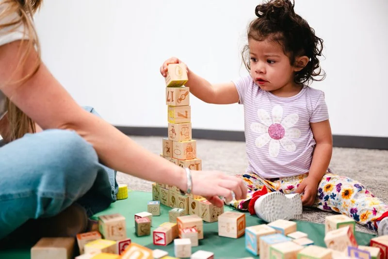 A young girl stacking wooden alphabet blocks on a mat while sitting on the floor, with other blocks scattered around her. An adult's arm is visible assisting.