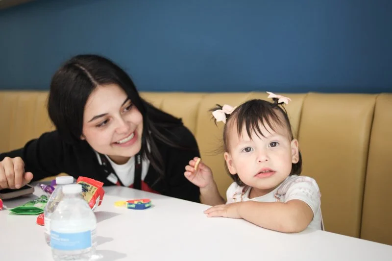 A woman and a young girl sitting at a table with toys, a water bottle, and snacks, in a restaurant or cafe setting.