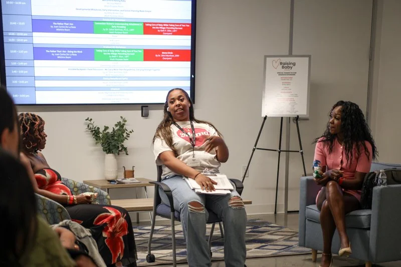 A woman is speaking during a panel discussion in a conference room, surrounded by three other women. The background features a large screen displaying event information and a poster on a stand. The women are seated on chairs with a small table and a 