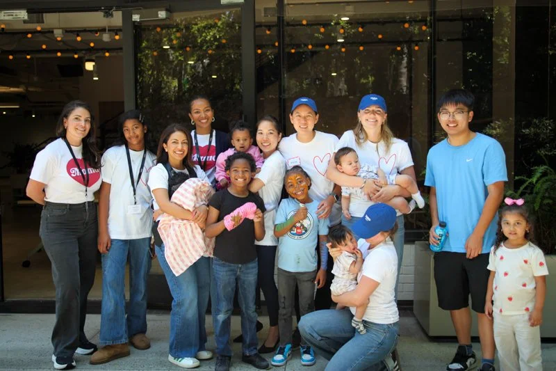 Group of diverse children and adults smiling, standing outside a building with glass doors, some holding babies and wearing casual clothes.