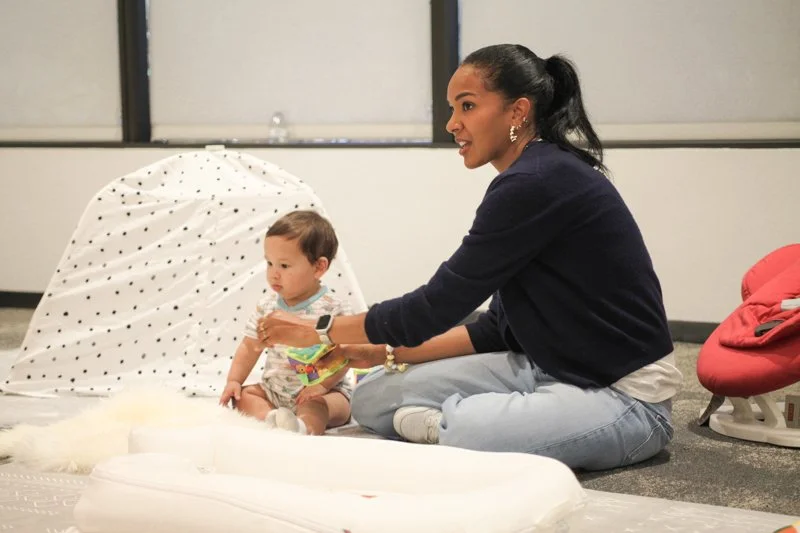 A woman sitting on the floor playing with a young child in a room with large windows, a polka-dotted tent, and a red baby rocker.