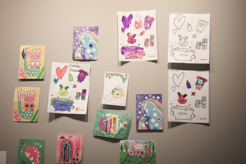 Collection of children's drawings and colorful pictures displayed on a wall, featuring animals, rainbows, hearts, and creative doodles.