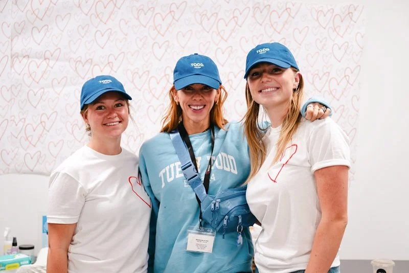 Three women smiling and wearing blue hats, two women wearing white t-shirts with red hearts, standing close together in front of a wall with pink heart drawings. The woman in the middle has a lanyard and a blue bag.