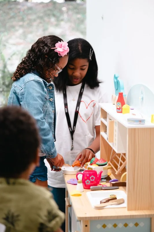 Two girls playing with toy food and kitchen set, while a boy watches, in a playroom.