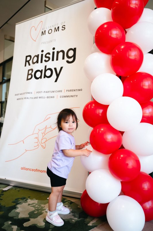 A young girl standing beside a large balloon display with red and white balloons at an event titled 'Raising Baby,' organized by Alliance of Moms.