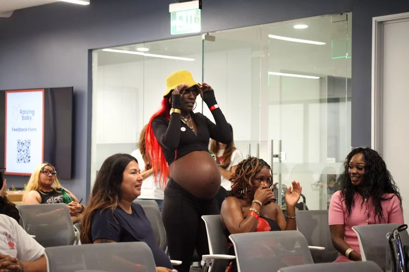 A pregnant woman standing and adjusting her yellow cap in a room filled with seated women. The women are smiling and observing her, and a glass wall and digital display are visible in the background.