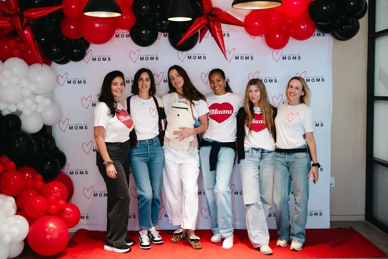 Six women standing together in front of a backdrop with balloons and the text "A Place of Moms," dressed casually with some wearing matching t-shirts, some with backpacks, smiling and posing for a photo.