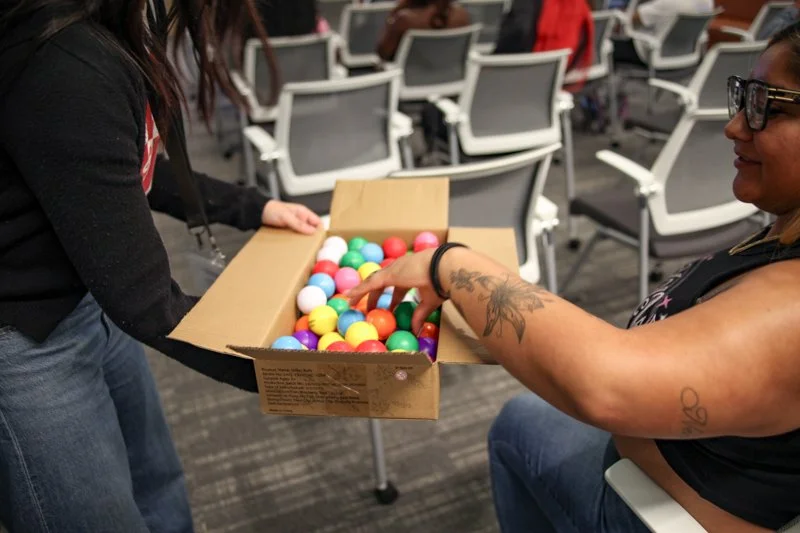 A woman with tattoos on her arm and glasses reaching for colorful gumballs from a cardboard box held by another person in an indoor setting with chairs and other people in the background.