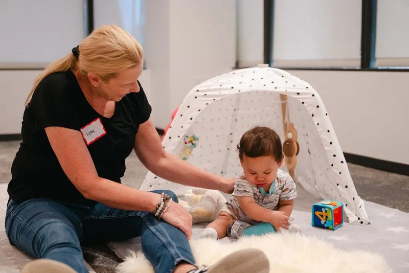 An older woman and a young boy playing on the floor with a small toy tent, a soft blanket, and a colorful cube in a modern room.