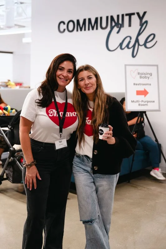 Two women standing in front of a sign that reads 'Community Cafe.' One woman is wearing a white shirt with a red heart and the word 'Mama,' and the other is in a black jacket. They are smiling and posing for the photo, with the woman in the black jac
