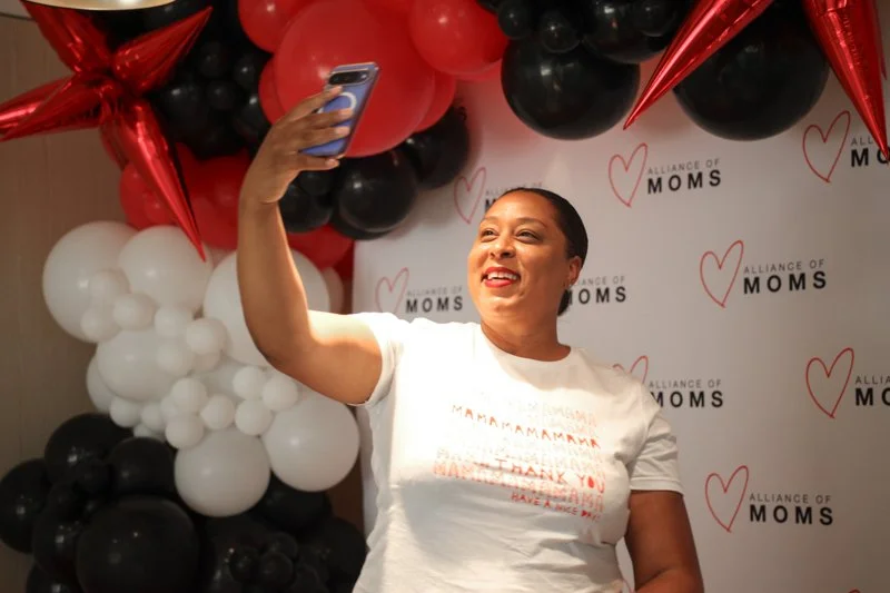 Woman taking a selfie in front of a decorated backdrop with balloons and "Alliance of Moms" logo.