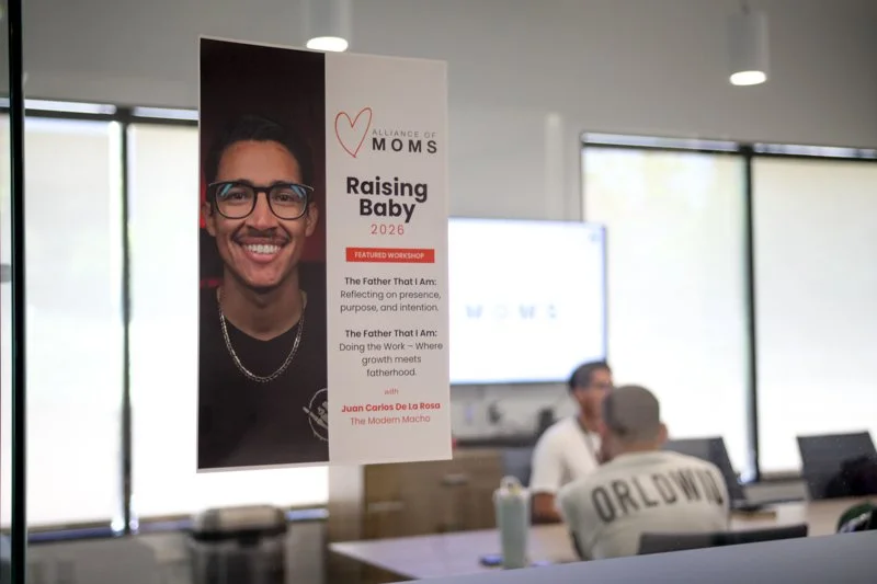 A vertical banner in a conference room promoting a workshop called 'Raising Baby 2026' by the Alliance of Moms, featuring a smiling man with glasses and a necklace. The banner includes details about the workshop, emphasizing reflection and growth in 