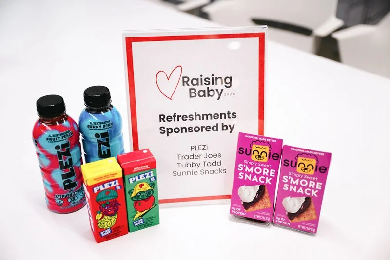 Assorted snacks and drinks on a table in front of a sign reading 'Raising Baby 2026, Refreshments Sponsored by, PLEZi, Trader Joes, Tubby Todd, Sunnie Snacks' at a baby-related event.