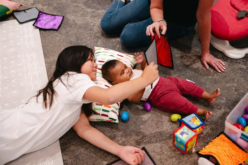 Two children, a girl and a toddler, lying on the carpeted floor playing with electronic tablets; the girl is smiling and resting her head on a pillow, while the toddler is lying next to her, looking at the tablet.