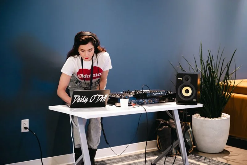 Young woman with headphones DJing at a white table with audio equipment, a laptop, and a speaker, in a room with a blue wall and a large potted plant.