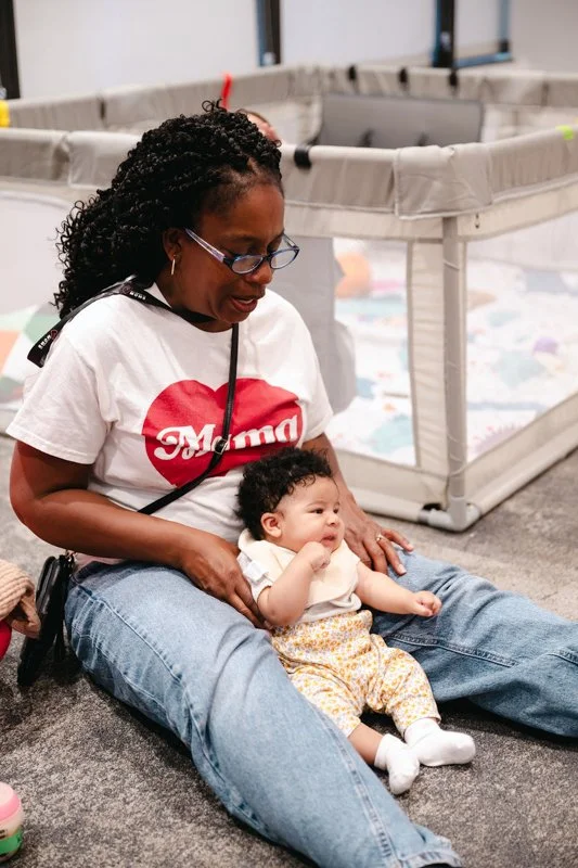A woman with glasses and curly hair sitting on the floor holding a baby with curly hair, in a room with cribs or playpens.