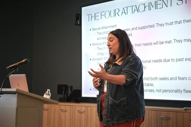 A woman giving a presentation in front of a large screen with the title "The Four Attachment Styles" displaying bullet points, in a classroom or conference room setting.