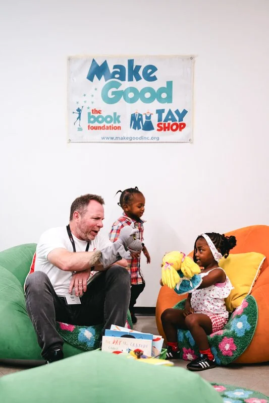 Two young girls, one sitting on an orange bean bag and the other standing, receiving a puppet from a smiling man with a beard. A banner reading 'Make Good' is on the wall above them.