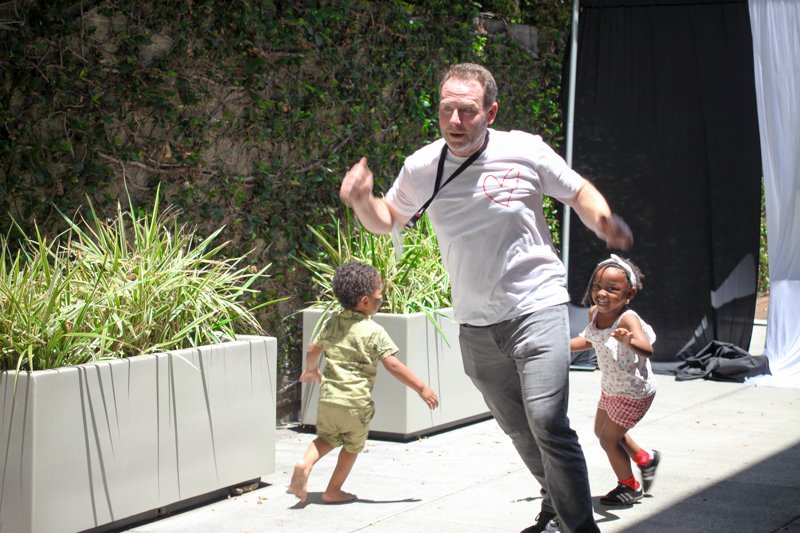 A man running outdoors with two children, one boy and one girl, in a garden-like area with large plants in white planters and a black backdrop.