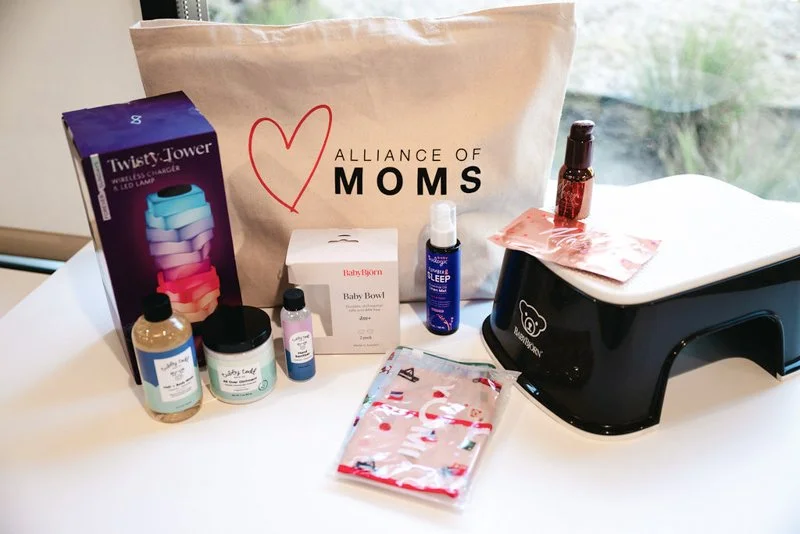 Collection of household items on a white table, including a TWIST and TOWER wireless charger, a beige tote bag with a red heart and the words 'ALLIANCE OF MOMS', a small white box labeled 'Baby Bowl', bottles of baby lotion, baby oil, a small bottle 