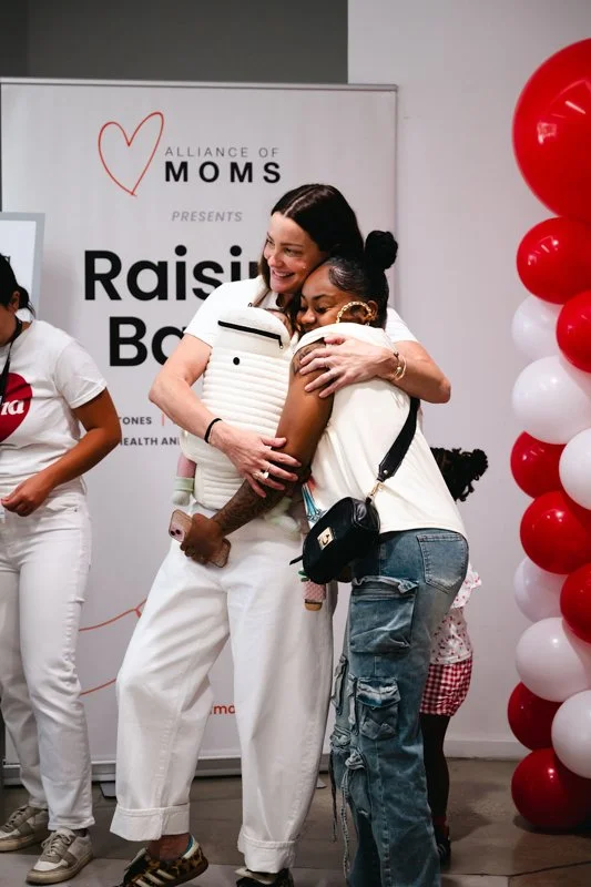 Two women hugging and smiling at a raising baby event, standing in front of a sign that reads "Alliance of Moms presents raising baby" with red and white balloons to the side.