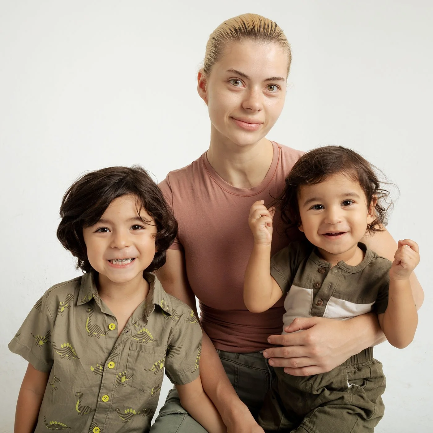A woman with blonde hair and two young children with dark brown hair, all smiling and looking at the camera, against a plain white background.