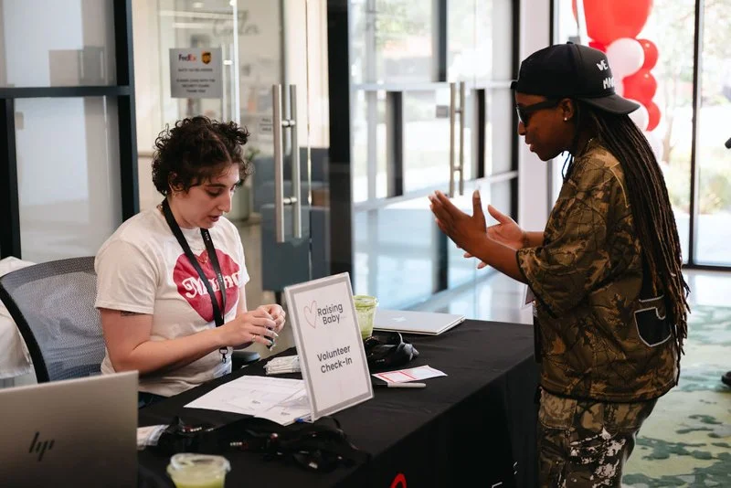 A woman with curly hair sitting at a volunteer check-in desk, speaking with a woman wearing sunglasses and a cap, in a modern indoor setting with large glass windows.