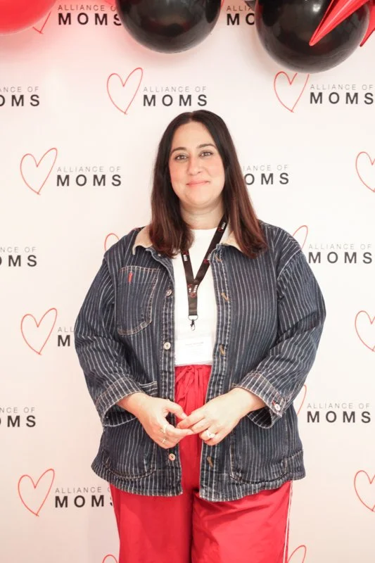 Woman standing in front of a backdrop with 'Alliance of Moms' written and heart graphics, wearing a striped jacket, red pants, and a lanyard around her neck.