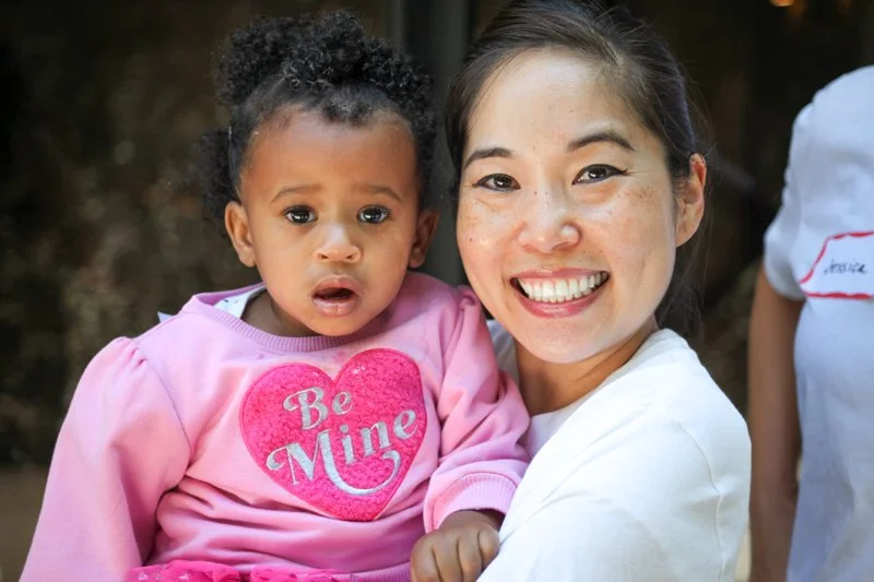 A smiling woman holding a young girl with a surprised expression, both outdoors. The girl wears a pink shirt with a heart that says 'Be Mine'.