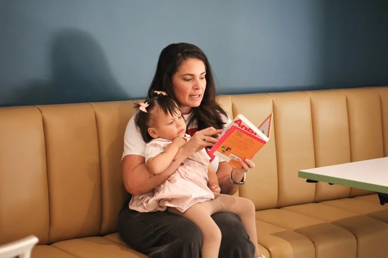 A woman sitting on a tan booth seat reading a book with a young girl sitting on her lap, in a setting that looks like a restaurant or cafe.