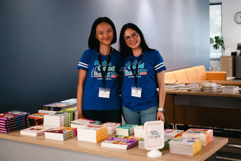 Two young women wearing blue T-shirts and nametags standing behind a table filled with colorful books at an indoor event.