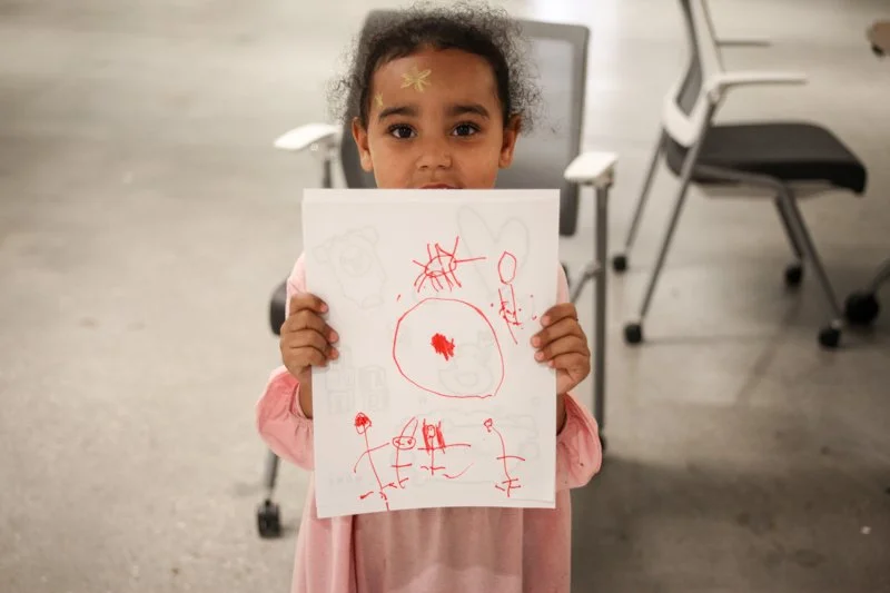 A young girl with curly hair, holding a piece of paper with red and gray crayon drawings, standing in a room with chairs and a gray floor.