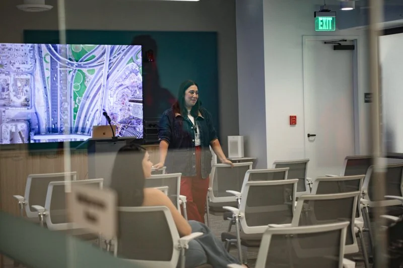 A woman standing at the front of a conference room with several empty chairs, a large screen displaying a cityscape, and an illuminated exit sign behind her.