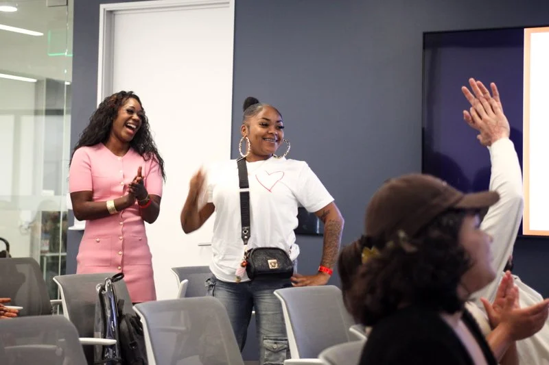 Two women standing and smiling during a meeting or presentation in a conference room, with one woman raising her hand and another clapping.