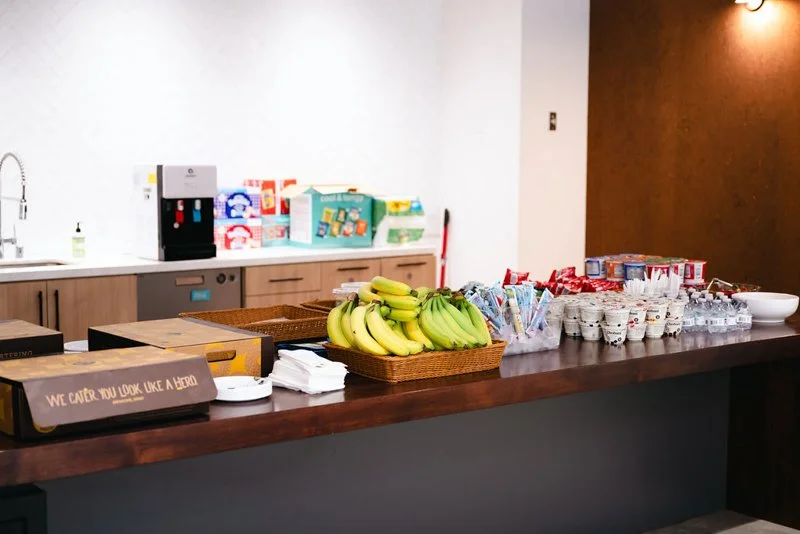 Snack bar with bananas, yogurt, water bottles, and cereal boxes on a counter in a break room or lounge area.