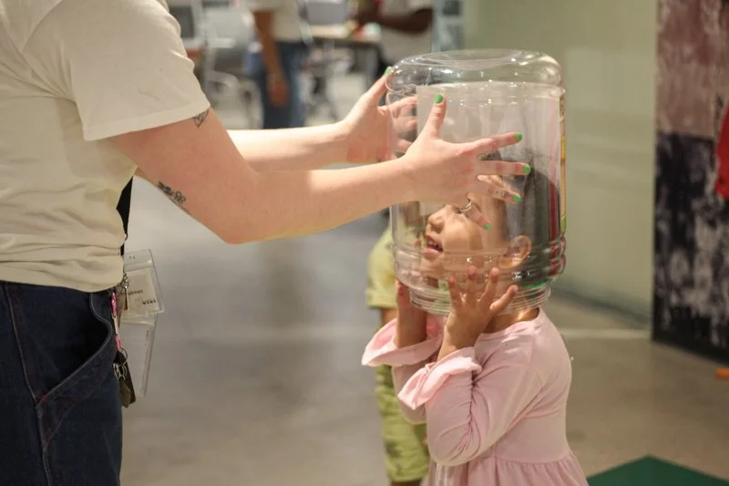 Child holding a large water jug over her head while an adult helps support it.