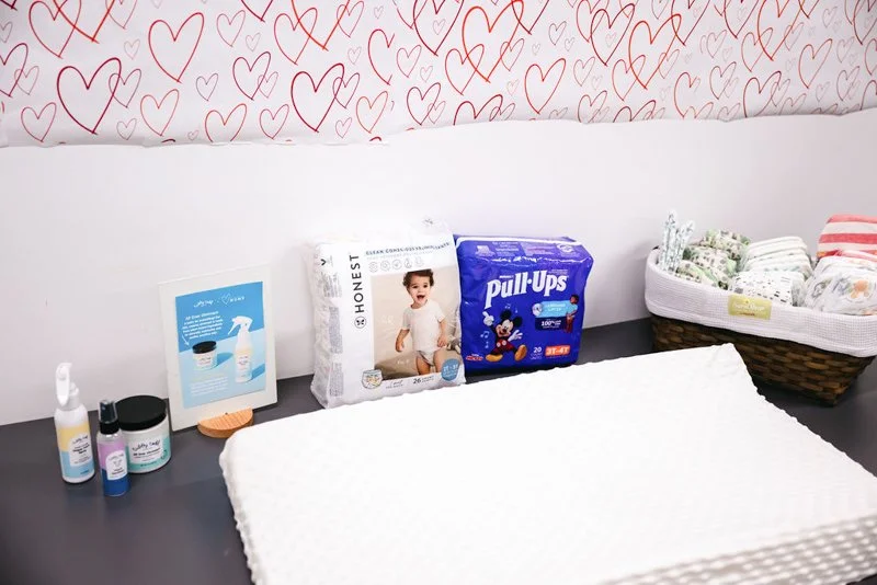 Baby diaper changing supplies on a dark changing table, including a pack of Honest diapers, a box of Pull-Ups training pants, bottles of diaper rash ointment, a framed photo or instructions, a small wooden brush, and a basket of rolled-up cloth diape