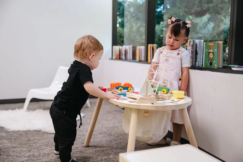 A young boy and girl playing with wooden toys at a table in a room with a bookshelf near a window.